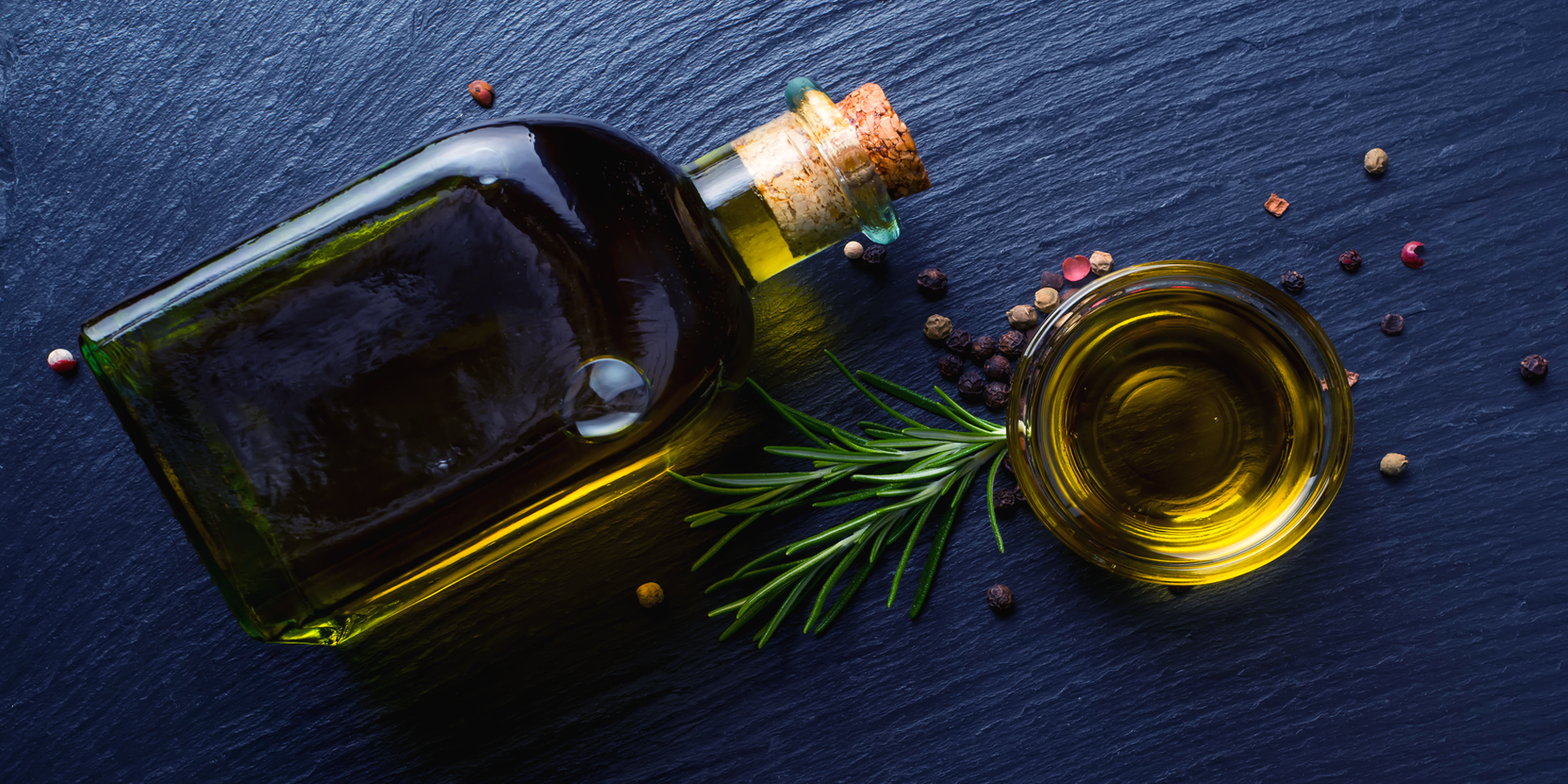 Lying bottle of olive oil with a cork, glass of olive oil, grains of pepper and a coniferous twig on the black surface